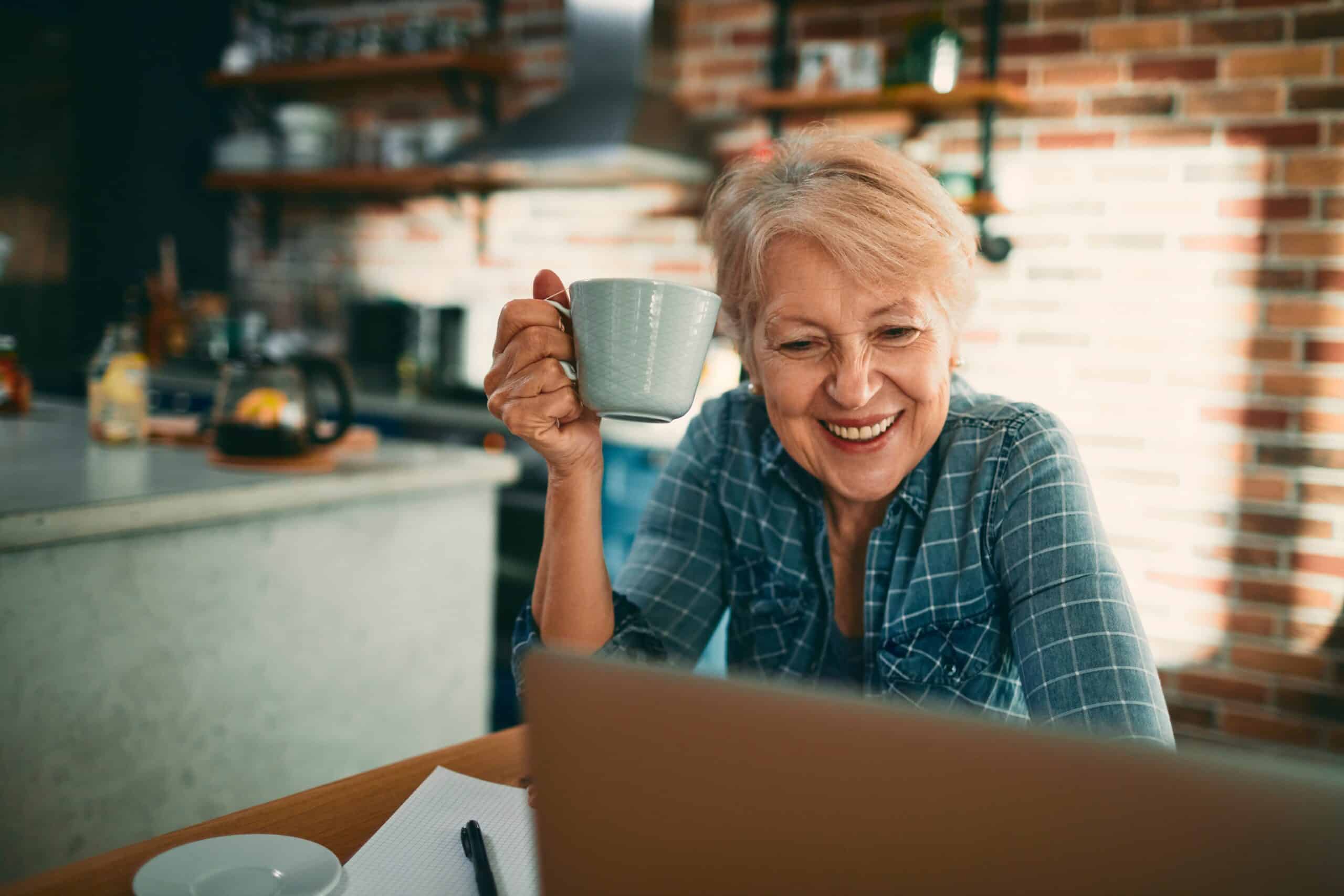 Smiling,Senior,Woman,Drinking,Coffee,And,Using,Laptop,At,Home
